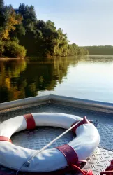 Ein Boot mit einem Rettungsring schwimmt auf einem ruhigen See. Im Hintergrund sind grüne Bäume und ein klarer Himmel sichtbar.