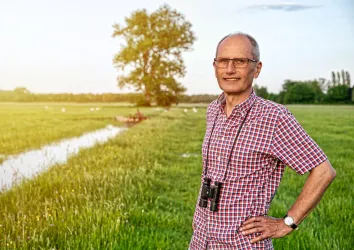 Ein Mann steht auf einer Wiese, neben ihm sind Bäume und ein Wasserlauf sichtbar. Er trägt ein kariertes Hemd und hat ein Fernglas um den Hals.