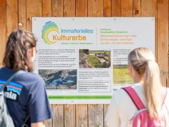 Zwei Personen stehen vor einem Informationsschild über immaterielles Kulturerbe. Das Schild enthält Texte und Bilder zu kulturellen Erbe-Stätten.