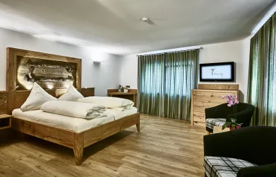 Ein modernes Schlafzimmer mit einem großen Bett und Holzdetails. Die Fenster sind mit grünen Vorhängen bedeckt und es gibt einen Fernseher sowie einen kleinen Tisch.