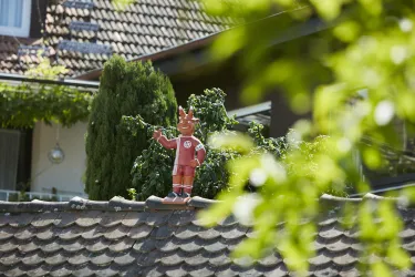 Eine rote Figur mit Hörnern steht auf einem Dach und zeigt mit dem Finger. Im Hintergrund sind grüne Pflanzen und ein sonniger Himmel zu sehen.