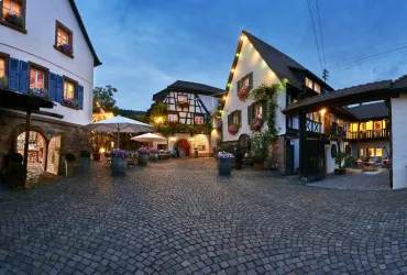 Ein malerischer Dorfplatz mit historischen Fachwerkhäusern und gemütlichen Sitzbereichen. Die Beleuchtung sorgt für eine einladende Atmosphäre am Abend.