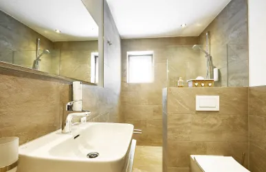 Ein modernes Badezimmer mit braunen Fliesen und großen Spiegeln. Die Ausstattung beinhaltet ein Waschbecken, eine Dusche und eine Toilette.