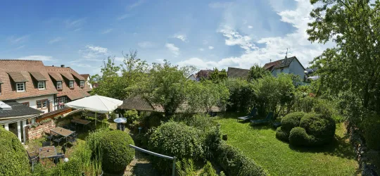 Ein gepflegter Garten mit Grünflächen und schattenspendenden Bäumen. Im Hintergrund sind mehrere Häuser und ein blauer Himmel zu sehen.
