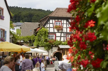 Ein malerisches Dorf mit Fachwerkhäusern und bunten Blumen. Menschen genießen die Atmosphäre in einem einladenden Ambiente.