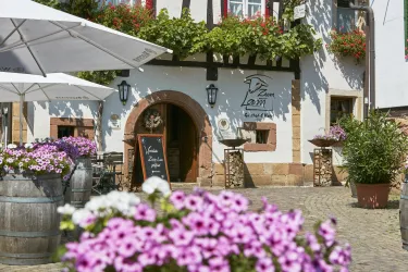 Ein malerisches Gasthaus mit einer einladenden Terrasse und blühenden Blumen. Der Platz ist sonnig und freundlich gestaltet.