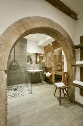 Ein modernes Badezimmer mit einem halbrunden Steinbogen. Es enthält eine Glasdusche, ein Waschbecken und Holzregale mit Handtüchern.