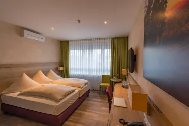 Ein modernes Hotelzimmer mit einem Doppelbett und großen, hellen Fenstern. Die Einrichtung ist schlicht und gemütlich, ergänzt durch einen Schreibtisch und einen Fernseher.