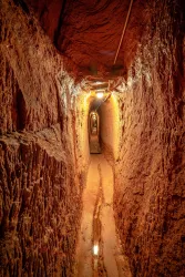 Ein schmaler Tunnel mit erdigen Wänden und einem leicht feuchten Boden. Über dem Tunnel verlaufen elektrische Leitungen und schwaches Licht beleuchtet den Weg.