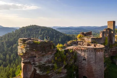 Eine beeindruckende Burgruine auf einem Felsen, umgeben von bewaldeten Hügeln. Im Hintergrund erstreckt sich eine malerische Landschaft unter blauem Himmel.