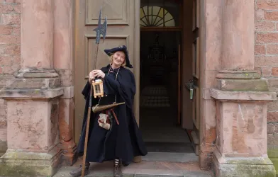 Eine Person in historischer Kleidung steht vor einer großen holzernen Tür. Sie hält eine Laterne und einen langen Stock in der Hand.