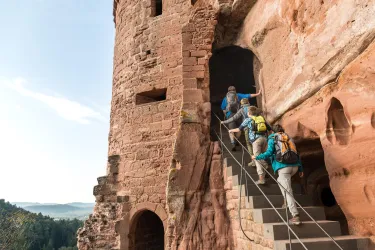 Eine Gruppe von Wanderern geht eine Treppe zu einem alten Turm hinauf. Die Umgebung ist hügelig mit einem klaren Himmel im Hintergrund.