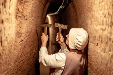 Eine Person arbeitet mit Werkzeugen in einem engen Tunnel. Die Wände sind aus braunem, bearbeitetem Material.