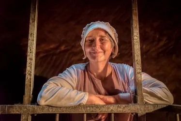 Eine lächelnde Frau blickt durch die Stäbe eines Gitters. Sie trägt eine Haube und historische Kleidung.