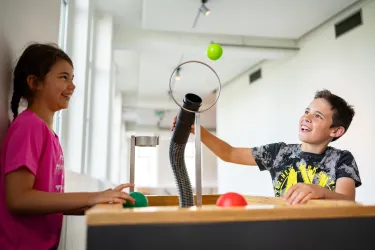 Zwei Kinder spielen an einem interaktiven Tischspiel. Ein Junge wirft einen grünen Ball durch einen ringförmigen Schlauch, während ein Mädchen zuschaut und lacht. Foto: Dynamikum Science Center Pirmasens/Sebastian Fachenbach