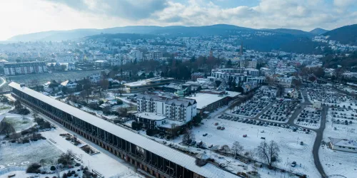 Eine winterliche Stadtansicht mit schneebedeckten Dächern und Berghintergrund. Im Vordergrund sind Wohngebäude und Parkplätze zu sehen.