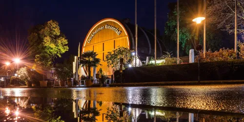 Ein großes, rundes Gebäude mit der Aufschrift "Dürheimer Riesenrad" ist zwischen Bäumen zu sehen. Im Hintergrund ist ein Uhrturm und weitere Gebäude zu erkennen.