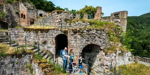 Eine Familie erkundet die Ruinen einer alten Burg. Sie stehen auf einem Weg umgeben von grüner Natur und historischen Steinen.