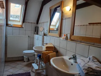 Ein modernes Badezimmer mit zwei Waschbecken und einem WC. Die Wände sind gefliest und es gibt viel Tageslicht durch die Dachfenster.