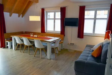 Ein helles Wohnzimmer mit einem großen Esstisch und modernen Stühlen. Die Wände sind in warmen Farbtönen gehalten, und es gibt große Fenster mit roten Vorhängen.