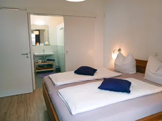 Ein gemütliches Schlafzimmer mit zwei Betten, eleganten Kissen und warmen Licht. Im Hintergrund ist ein modernes Badezimmer sichtbar.