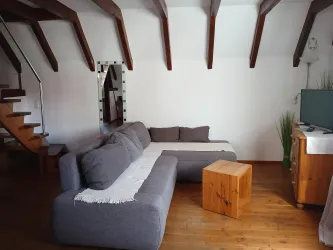 Ein gemütliches Wohnzimmer mit einem grauen Sofa und einem Holztisch. Die Holzdecke und die Treppe verleihen einen rustikalen Charme.
