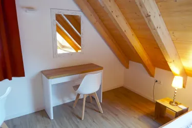 Ein gemütliches Zimmer mit schrägen Holzdecken und einem großen Spiegel. Der Raum ist mit einem Schreibtisch, einem Stuhl und einer kleinen Lampe ausgestattet.