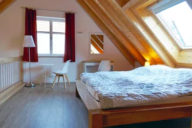Ein gemütliches Schlafzimmer mit Holzdecke und großen Fenstern. Es gibt ein großes Bett, einen Tisch und einen Stuhl sowie eine Stehlampe.