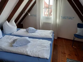 Ein gemütliches Schlafzimmer mit zwei Betten und Holzdecken. Es gibt einen Sessel und ein Fenster mit Vorhängen, das viel Licht hereinlässt.