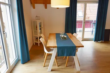 Ein einladender Essbereich mit einem Holztisch und weißen Stühlen. Große Fenster sorgen für viel Tageslicht und eine gemütliche Atmosphäre.