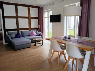 Ein modernes Wohnzimmer mit einem grauen Sofa und einem Essbereich. Große Fenster lassen viel Tageslicht herein.