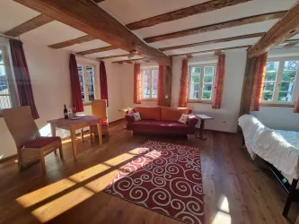 Ein gemütlicher Wohnraum mit Holzbalken und großen Fenstern. Die Einrichtung umfasst ein Sofa, einen Tisch und Stühle, sowie einen Teppich.