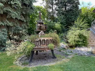 Eine skurrile Gartenfigur aus Metall, die mit Pflanzen dekoriert ist. Im Hintergrund sind Bäume und hohe Sträucher zu sehen.
