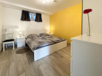 Ein gemütliches Schlafzimmer mit einem Doppelbett und modernen Möbeln. Die Wände sind in warmen Farben gestrichen, und eine Blume in einer Vase sorgt für einen einladenden Akzent.