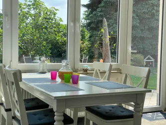 Ein helles Esszimmer mit einem weißen Tisch und bunten Gläsern. Durch die großen Fenster ist viel Tageslicht und der Blick auf den Garten ist sichtbar.