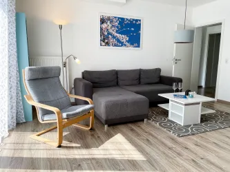 Ein helles Wohnzimmer mit einem grauen Sofa und einem modernen Sessel. Der Raum ist freundlich eingerichtet und hat einen schönen Blick auf ein blühendes Bild an der Wand.