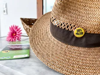 Ein sonniger Strohhut mit einem braunen Band und einem gelben Aufkleber mit der Aufschrift "100% PÄLZER". Neben dem Hut liegt eine pinke Blume und einige Informationsblätter.