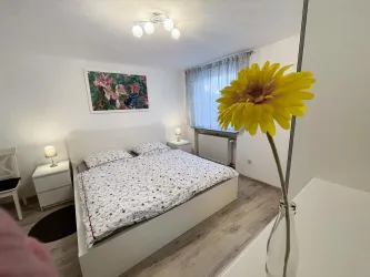Ein gemütliches Schlafzimmer mit einem großen Bett und bunten Kissen. Ein gelber Blumenstrauß steht auf dem Tisch und sorgt für einen frischen Akzent.