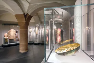 Ein modernes Museum mit ausgestellten Kunstgegenständen in Vitrinen. Im Vordergrund befindet sich eine dekorative Schüssel, umgeben von weiteren Artefakten.