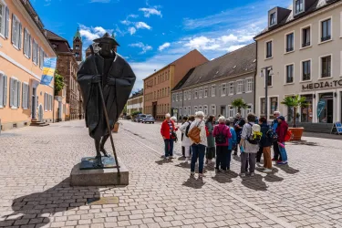 Eine Statue von einem Mann steht auf einem Platz, umgeben von historischen Gebäuden. Eine Gruppe von Touristen hört aufmerksam zu und betrachtet die Statue.