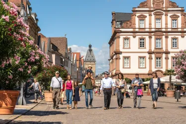 Eine belebte Straße mit Menschen, die fröhlich spazieren gehen. Im Hintergrund sind schöne Gebäude und blühende Pflanzen zu sehen.