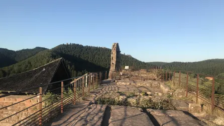 Eine historische Ruine auf einem Hügel mit Blick auf bewaldete Berge. Der Himmel ist klar und die Atmosphäre ist friedlich.