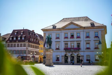 Ein schöner Platz mit einem historischen Gebäude und einer Statue. Die Umgebung ist von bunten Fenstern und grünen Pflanzen umgeben.