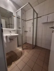 Ein modernes Badezimmer mit einer Dusche, einem Waschbecken und einem großen Spiegel. Die Wände sind hell gefliest und die Ausstattung ist sauber und funktional.