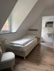 Ein einfaches Zimmer mit einem Einzelbett und einem Sessel. Im Hintergrund ist ein Badezimmer mit einem Waschbecken und einem Spiegel zu sehen.