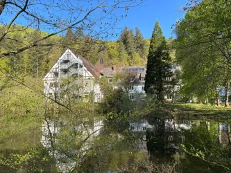 Ein malerisches Gebäude inmitten von grünem Wald und einem ruhigen Teich. Die Umgebung ist von Bäumen und frischem Blattwerk umgeben.