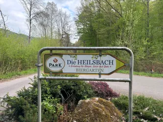 Ein Wegweiser zum Park "Die Heilsbach" mit Informationen über den Biergarten. Die Umgebung ist grün und naturnah.