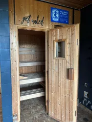 Eine Holzsauna mit offener Tür steht im Vordergrund. Der Schriftzug „get naked“ befindet sich über der Tür.