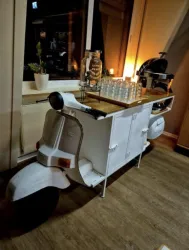 Ein weißes Motorrad mit einem Tisch darauf, der mit Gläsern und einem Getränkespender dekoriert ist. Der Hintergrund ist warm beleuchtet und vermittelt eine gemütliche Atmosphäre.