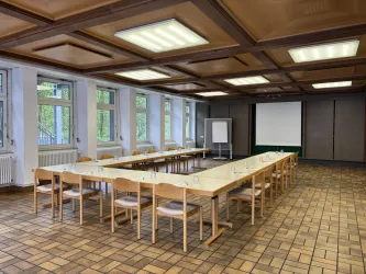 Ein großer Konferenzraum mit langen Tischen und Stühlen. Der Raum hat Fenster mit Blick nach draußen und eine Präsentationsfläche an der Wand.